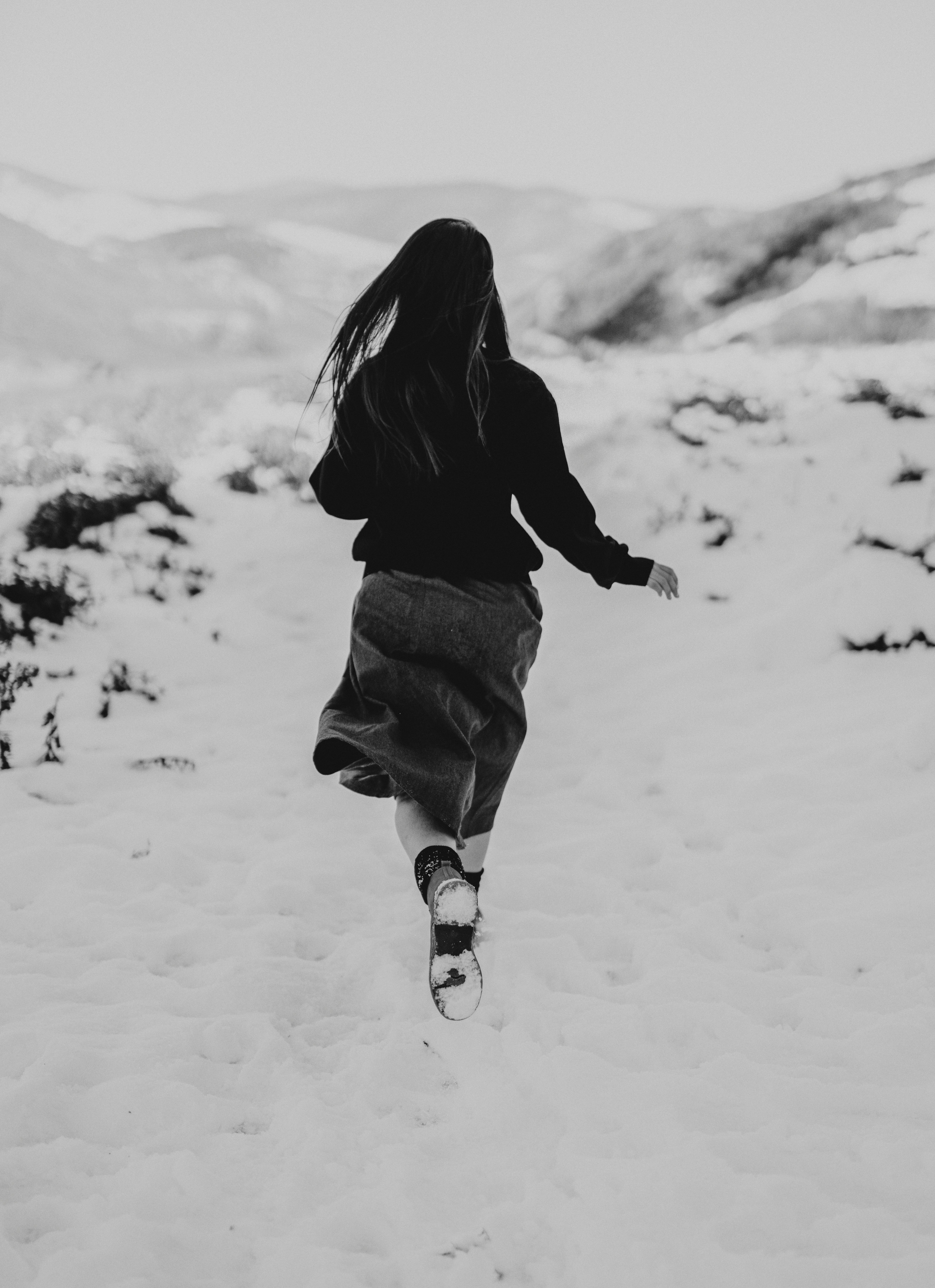 grayscale photo of woman in black hoodie and black skirt running on snow covered ground